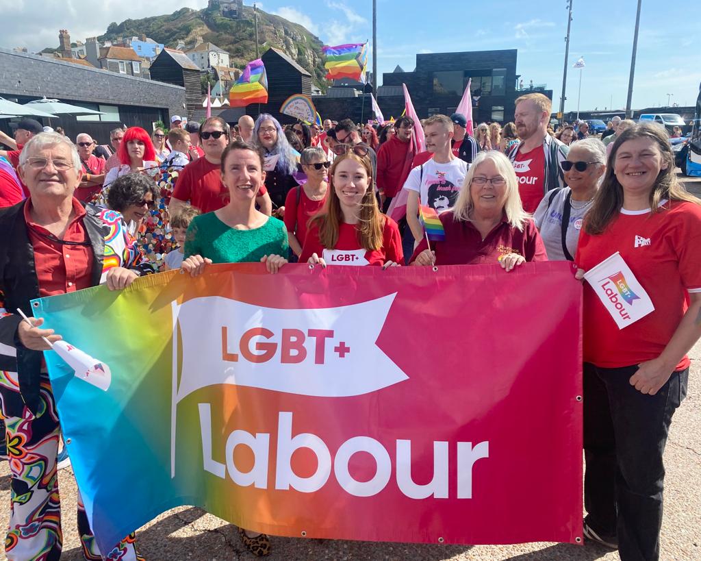 A group of smiling people, mostly dressed in red, standing outside and holding an "LGBT+ Labour banner".