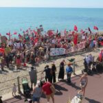 A large crowd of people on a beach, holding red flags and banners.